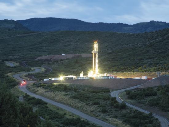 Mountain setting at dusk with a winding road and lighted tower showing oil and gas development.