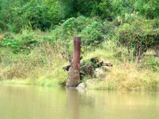 An orphan well with rusted equipment above surrounded by weeds next to water