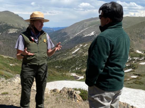 Man in green jacket speaks with park ranger with mountain scenery and blue sky in background.