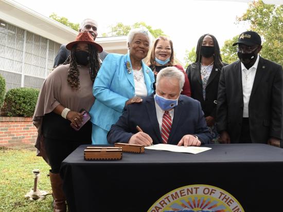 Secretary Bernhardt sits at a table in front of a brick house and signs a paper as a small group of people look on.