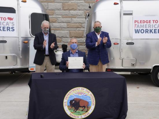 With two metal RVs in the background, Secretary Bernhardt looks on as a man sits at a table and holds up a piece of paper.