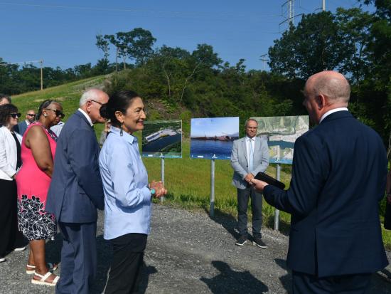 Secretary Haaland and group of people stand around 3 display panels during a site visit in NY. 