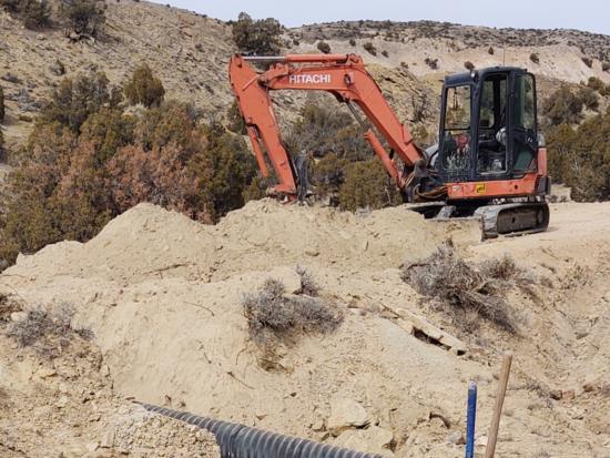 An excavator pulls up dirt, surrounded by dirt hills and shrubs.