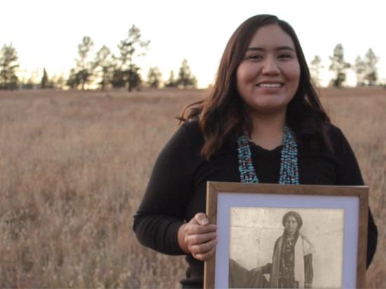An Indigenous woman holds up a framed black & white photograph of an ancestor 
