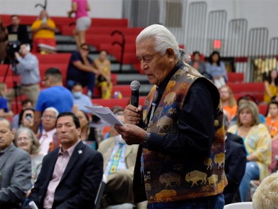 Older Native man speaks into microphone inside a crowded gymnasium 
