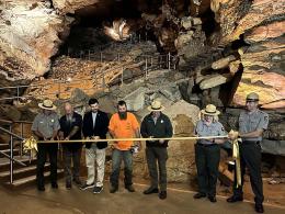 Seven people each cut a long yellow ribbon in front of a rocky hill inside a cave.
