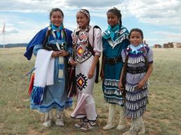 Four young girls in traditional Native American clothes pose together in grassy field.
