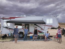 Campers stand in front of an RV in a sandy terrain