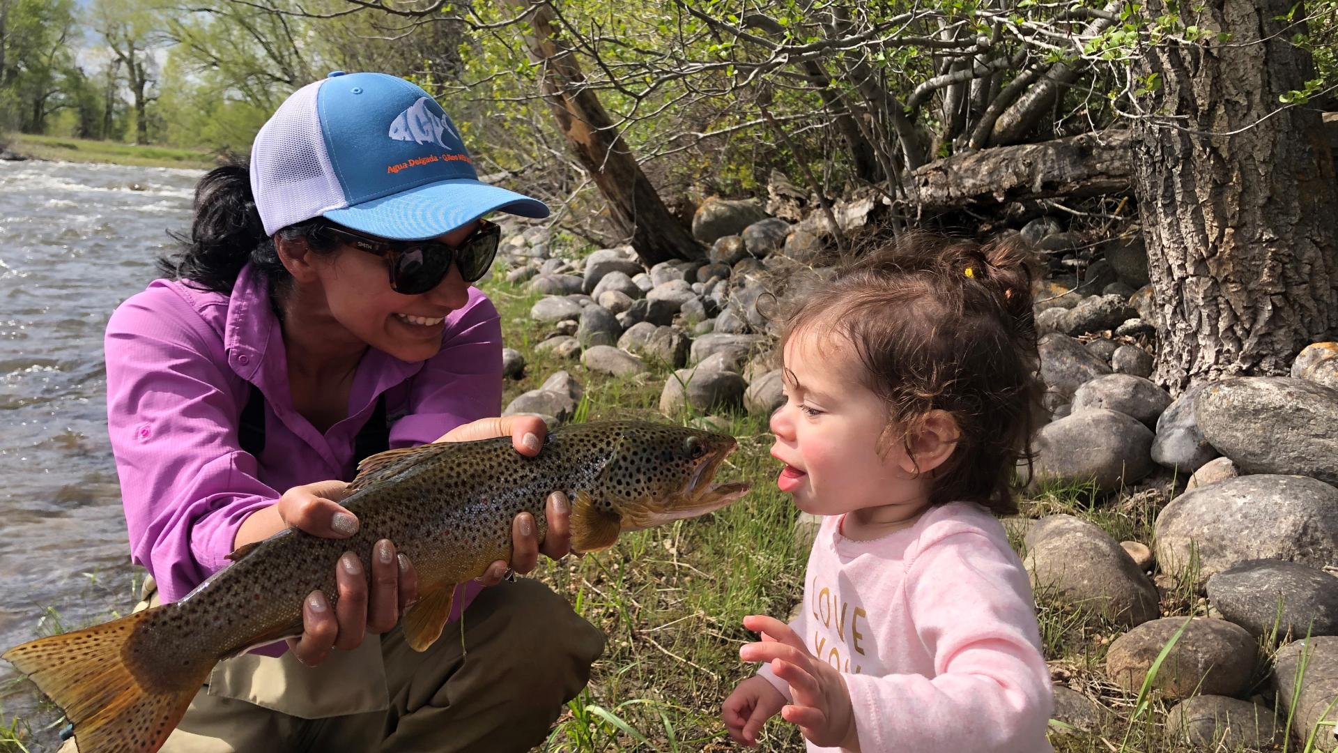 Karen shows her toddler a freshly caught trout along a river bank