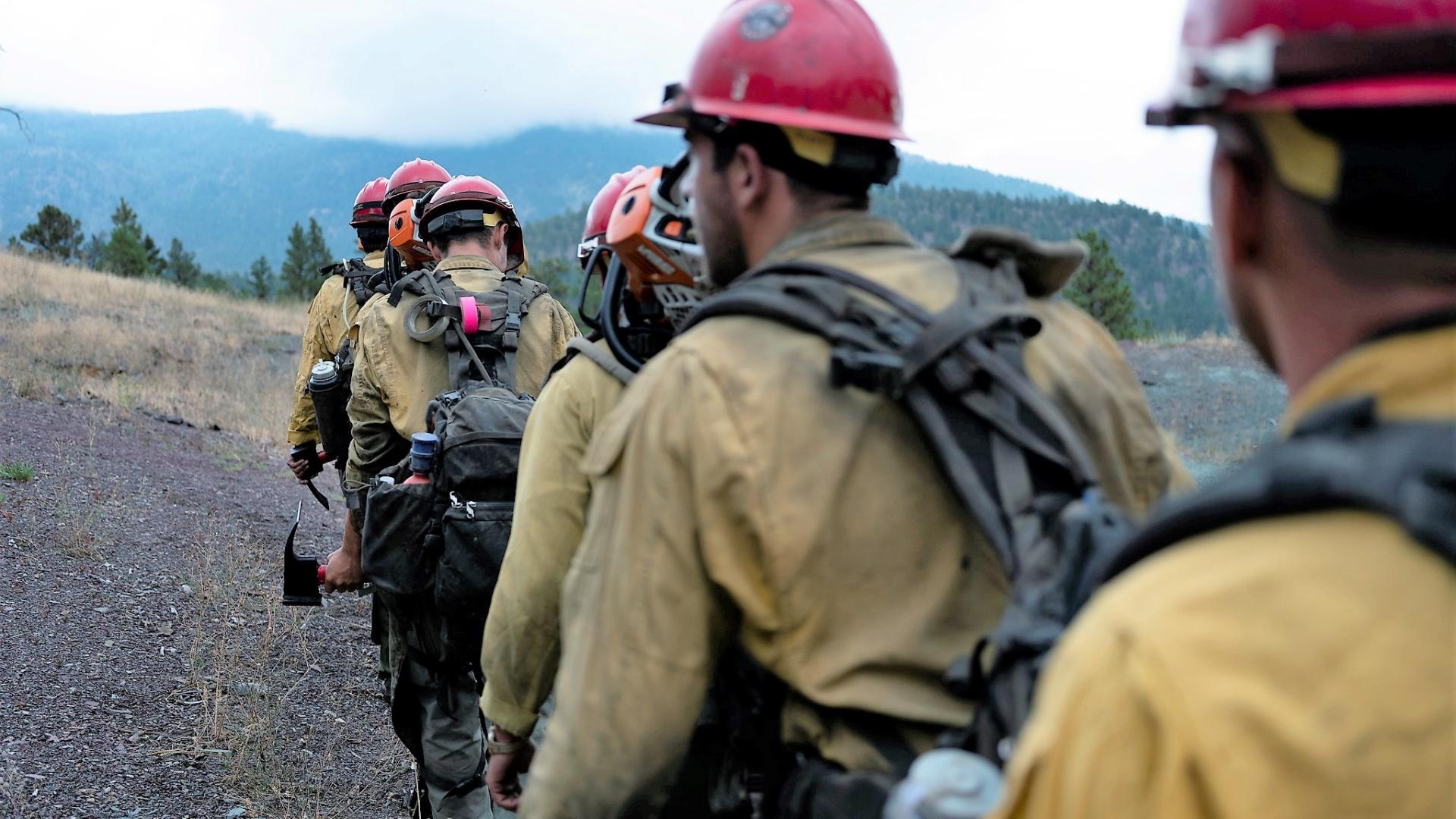 Wildland firefighters hike in a line wearing their fire gear and carrying tools. 