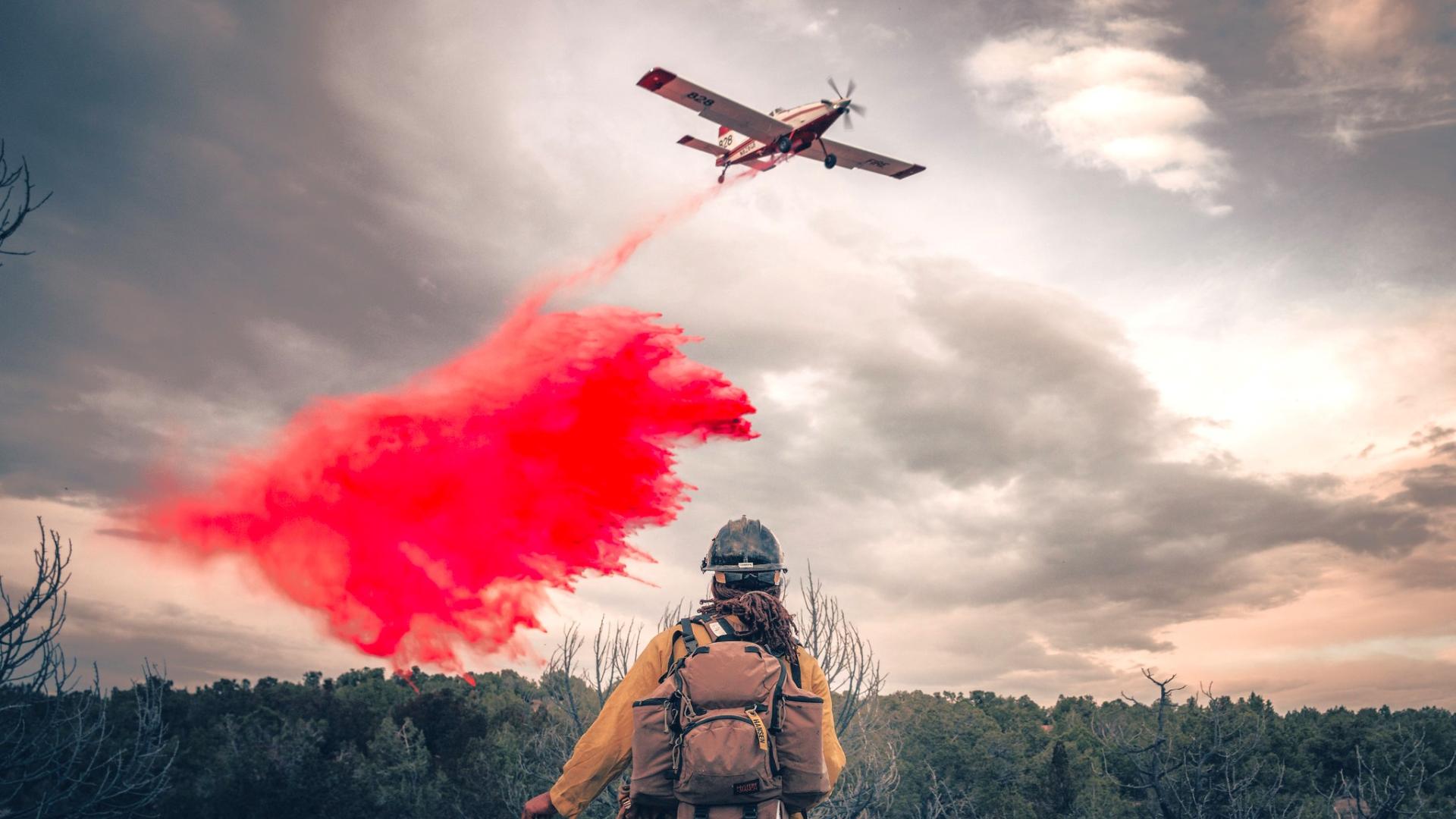 A wildland firefighter watches as a single engine air tanker drops red retardant on a wildfire. The firefighter's back is to the viewer as the plane flies overhead, with smoke and clouds in the background. BLM photo. 