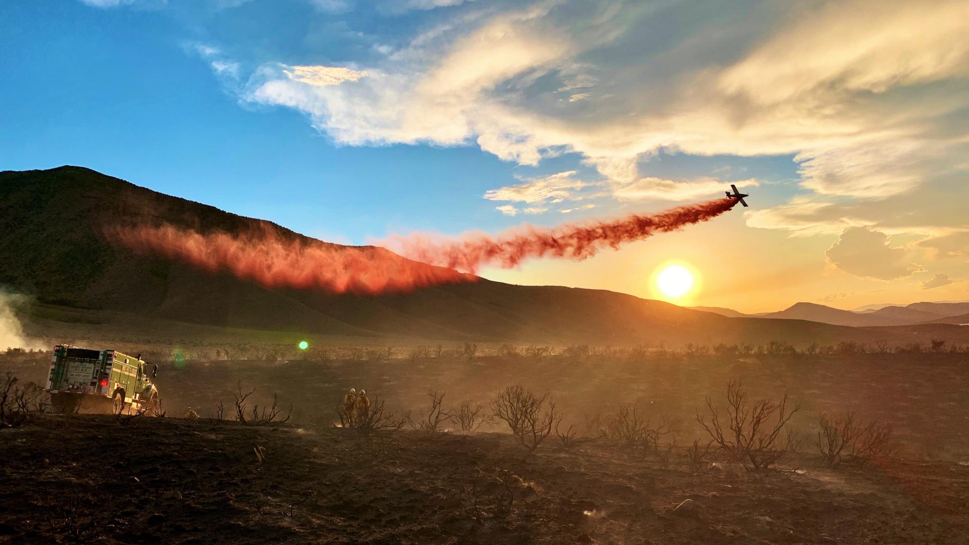 A single engine air tanker drops retardant on a wildfire. A red stream streaks the sky as the retardant exits the plane, with the sun setting in the background and burned vegetation in the foreground. BLM photo.