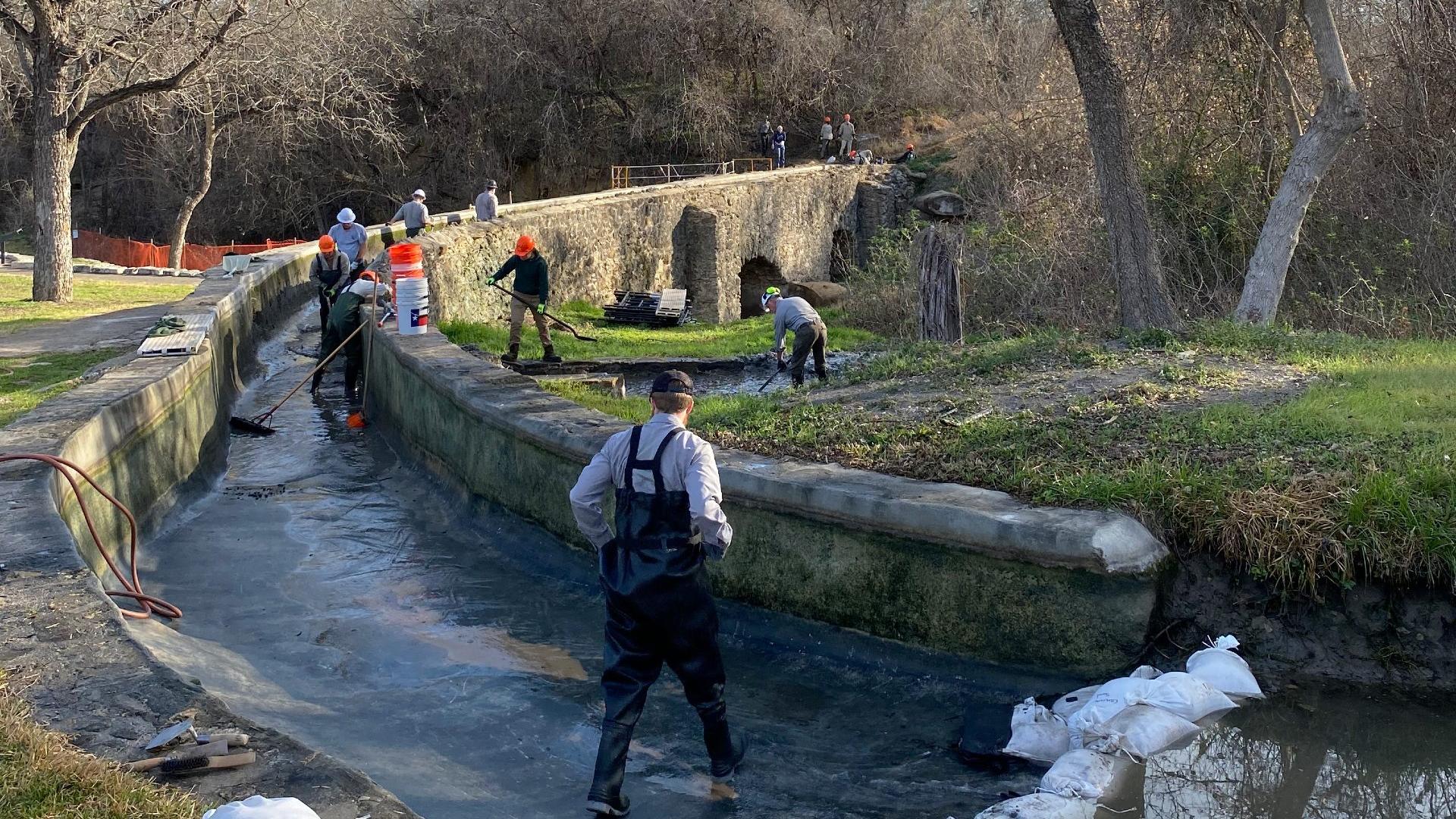 A group of eight construction workers repair a curved stone aqueduct surrounded by bare trees