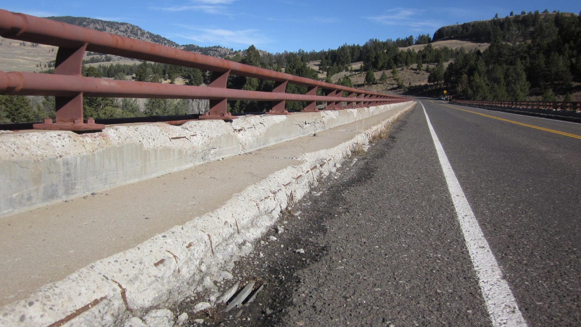 Close-up photo of a paved road with red railings with pine trees and hills in the distance.