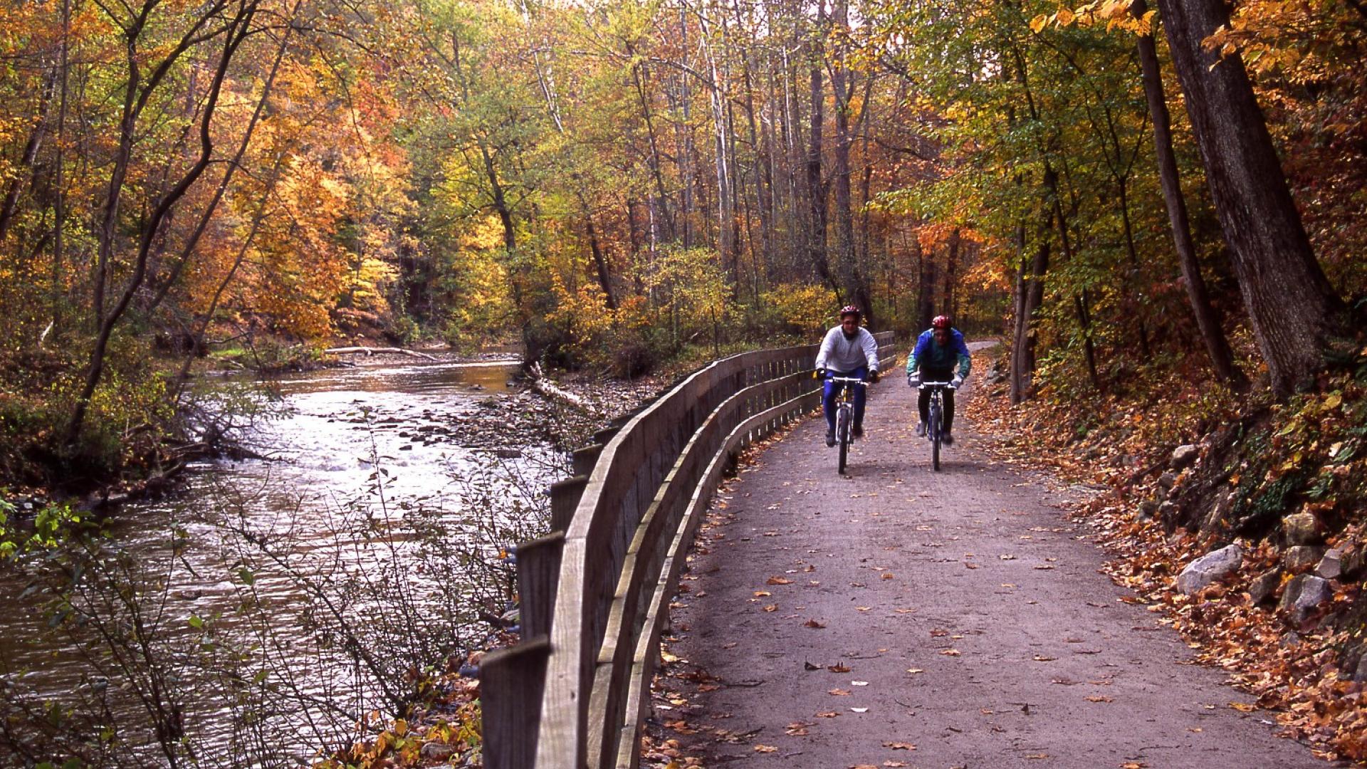 Two people on bicycles on a flat trail surrounded by fall forest colors. A wooden fence in the center divides the trail to the right and the river to the left.