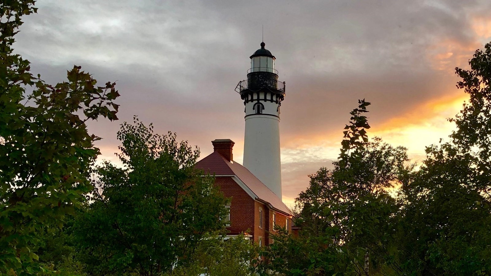 White lighthouse behind a red brick building at sunset.