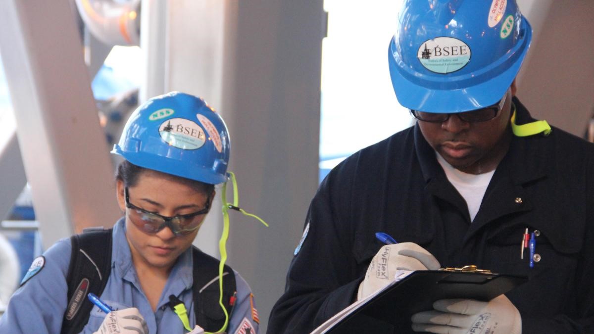 A man and a woman wearing hardhats review documents on a clipboard.