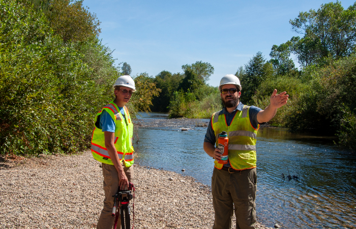 A couple of men wearing safety vests and white hardhats and standing by a river with trees on either side