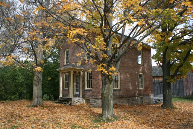 A small brick house with a porch surrounded by trees showing their autumn colors.
