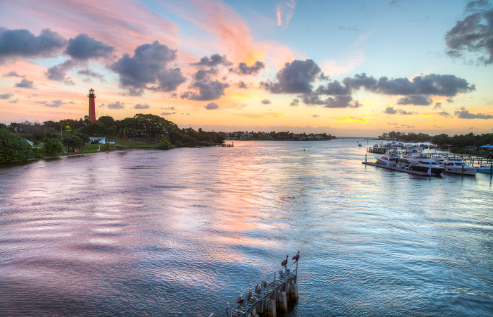 A blue and pink sunset reflects on a body of water with boats and trees around it and red lighthouse in distance