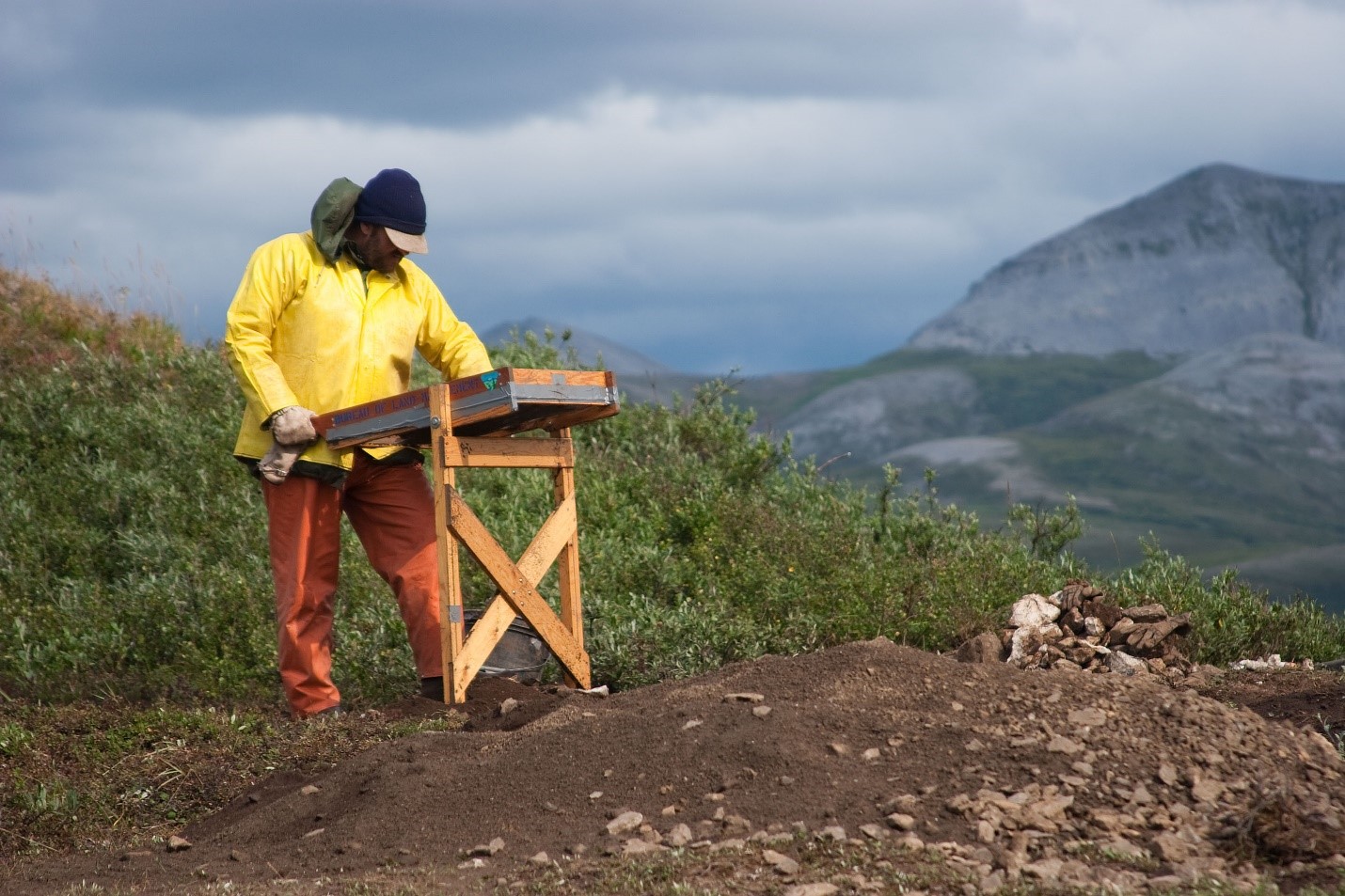 Man on a hilltop works behind a wooden table with mountains in the background.