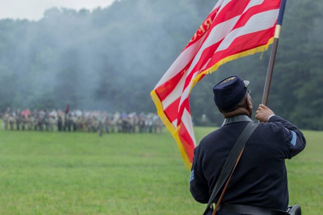 An African American man wearing a Union Army uniform and waving an American flag in a green field during a battle reenactment.