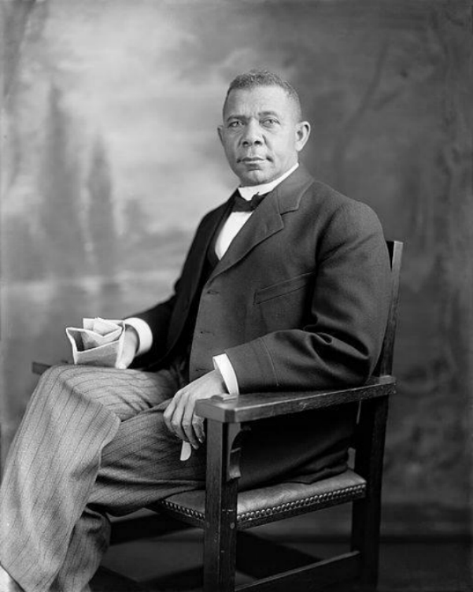 A historic black and white photo of a distinguished looking African American man in a suit sits in a wooden chair.