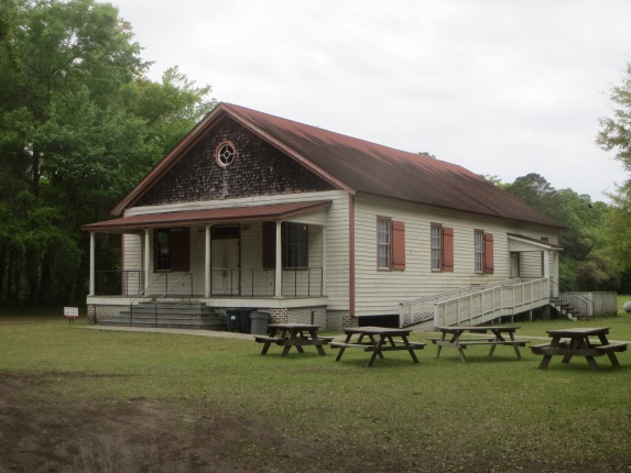 A long, one story wooden hall with a wide porch and picnic tables on the front lawn.