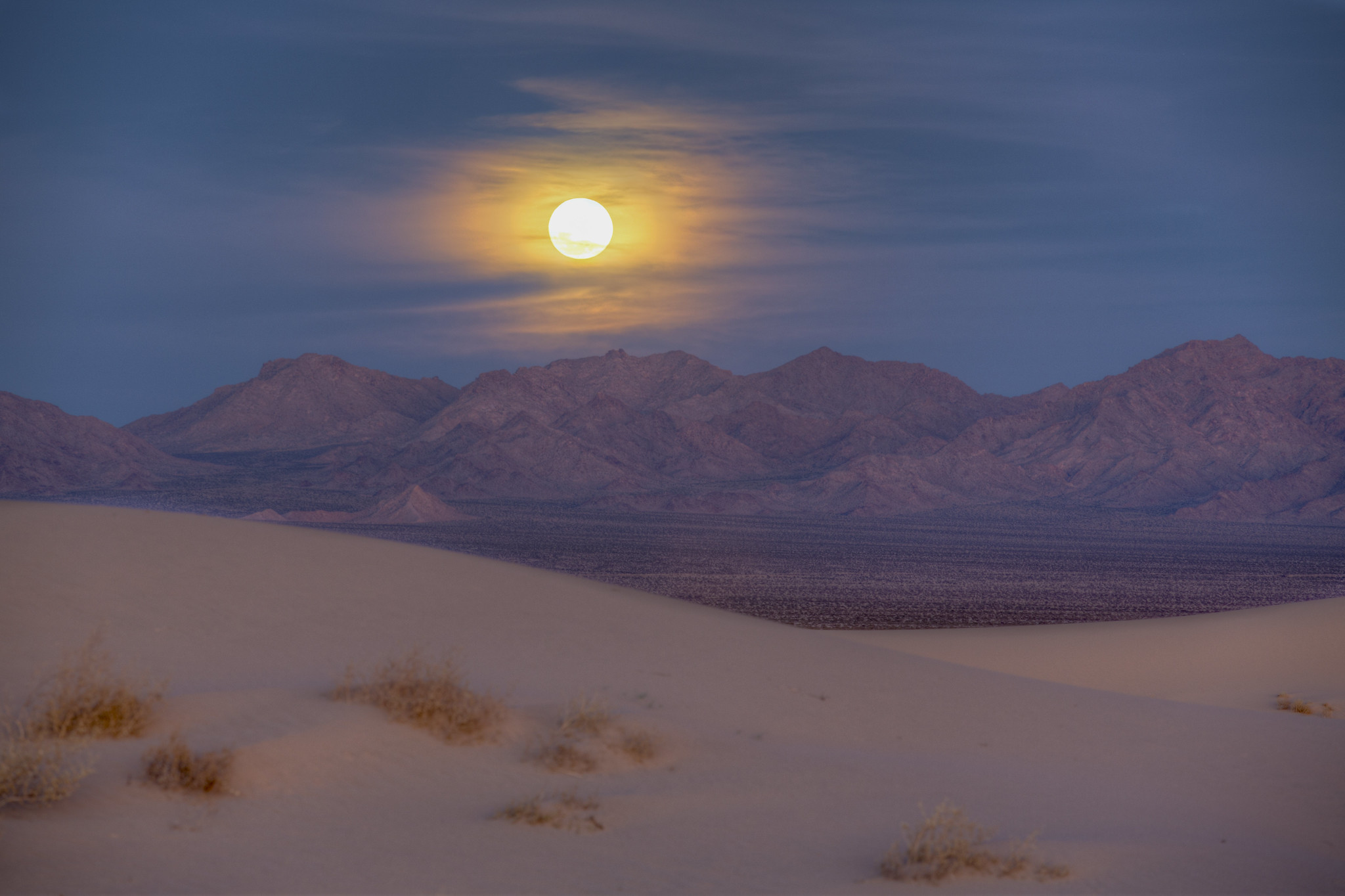 Cloudy sky with full moon over a purple mountain range and sand dunes.