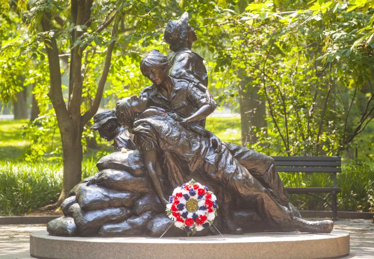 Brown statue of one woman sitting on a rock as a man lays in her arms, another woman looks towards the sky, and a final woman looks at the ground. Green trees are behind the statue and a red, white, and blue wreath sits in front of it. 