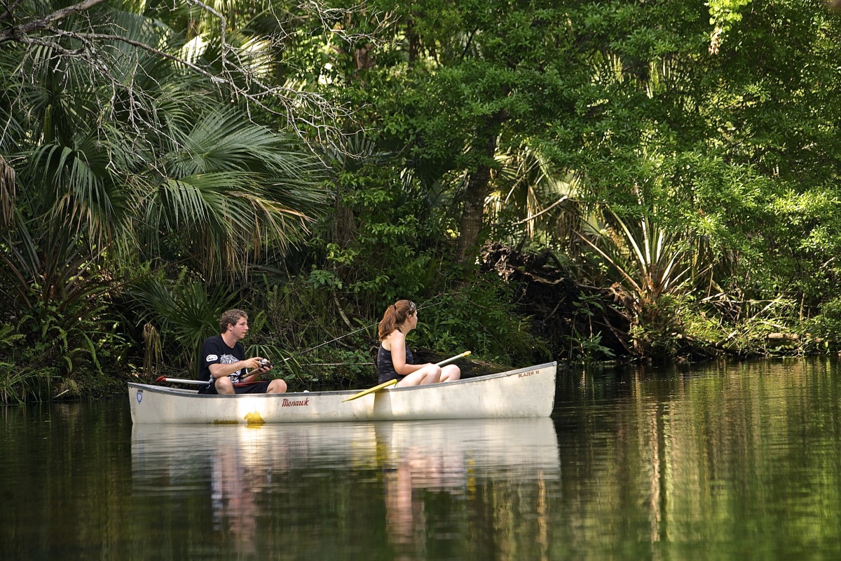A man and a woman sit in a canoe along the bank of the Wekiva River. The man fishes while the woman watches. 