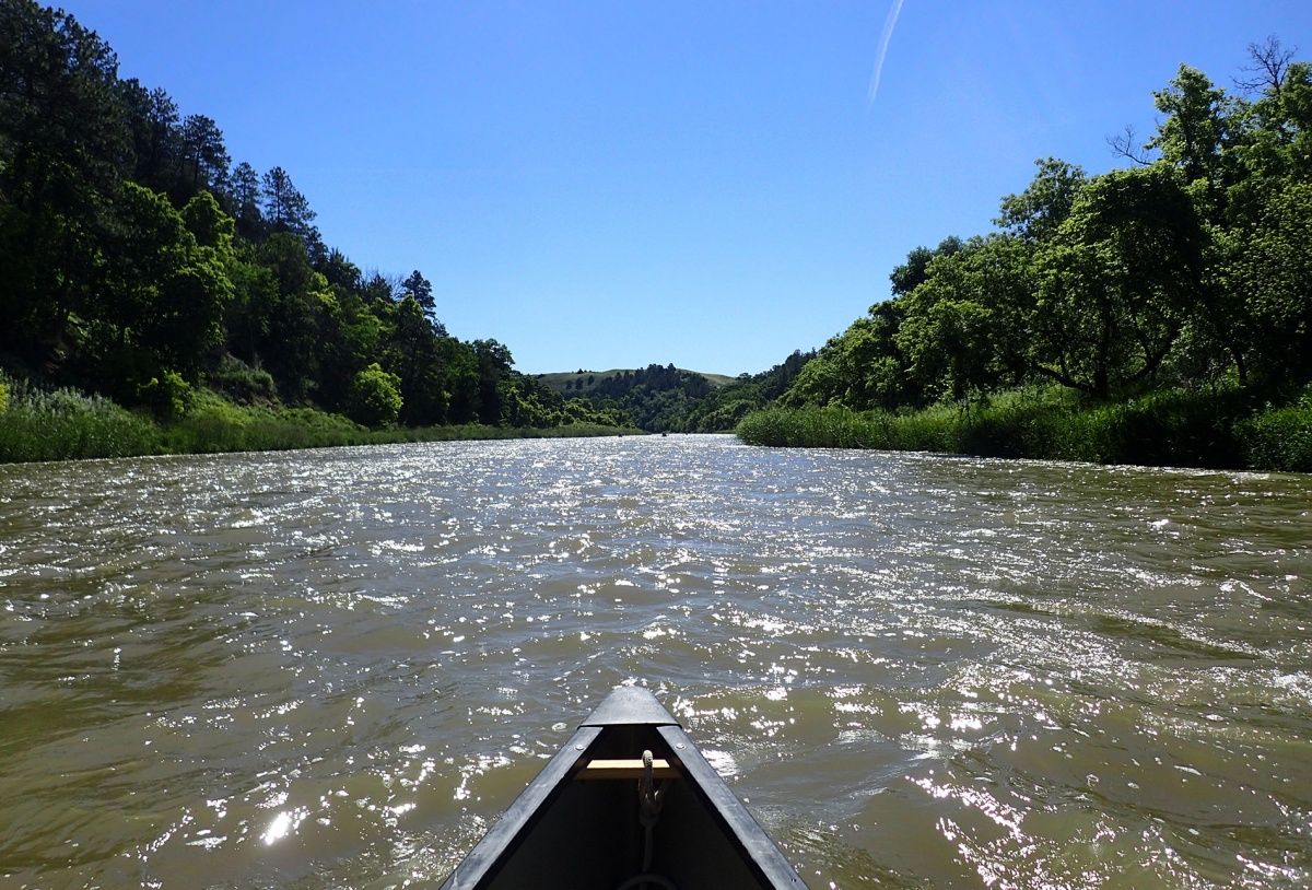 The front of a canoe sits at the bottom of the picture, showing a wide view of slow waves on the Niobrara River.