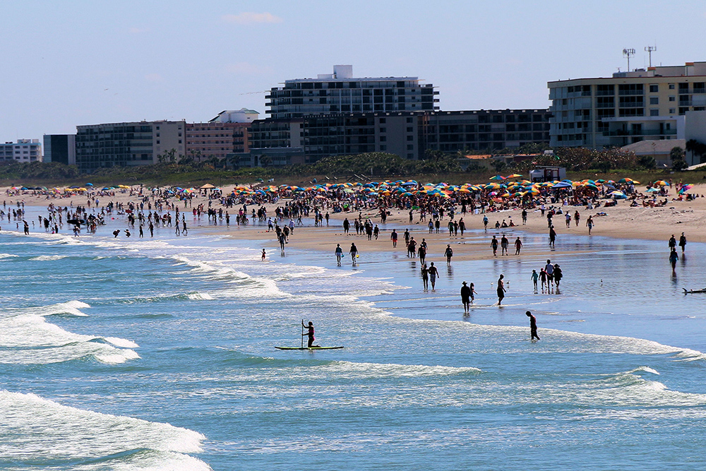 A beach is filled with people in the water and on the sand. Large buildings line the far side of the beach.