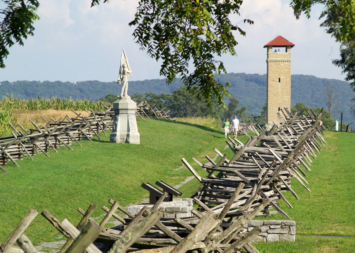 Wooden barriers line a pathway of green grass. A monument and a tower stand in the path, and a couple walks through the old battlefield.