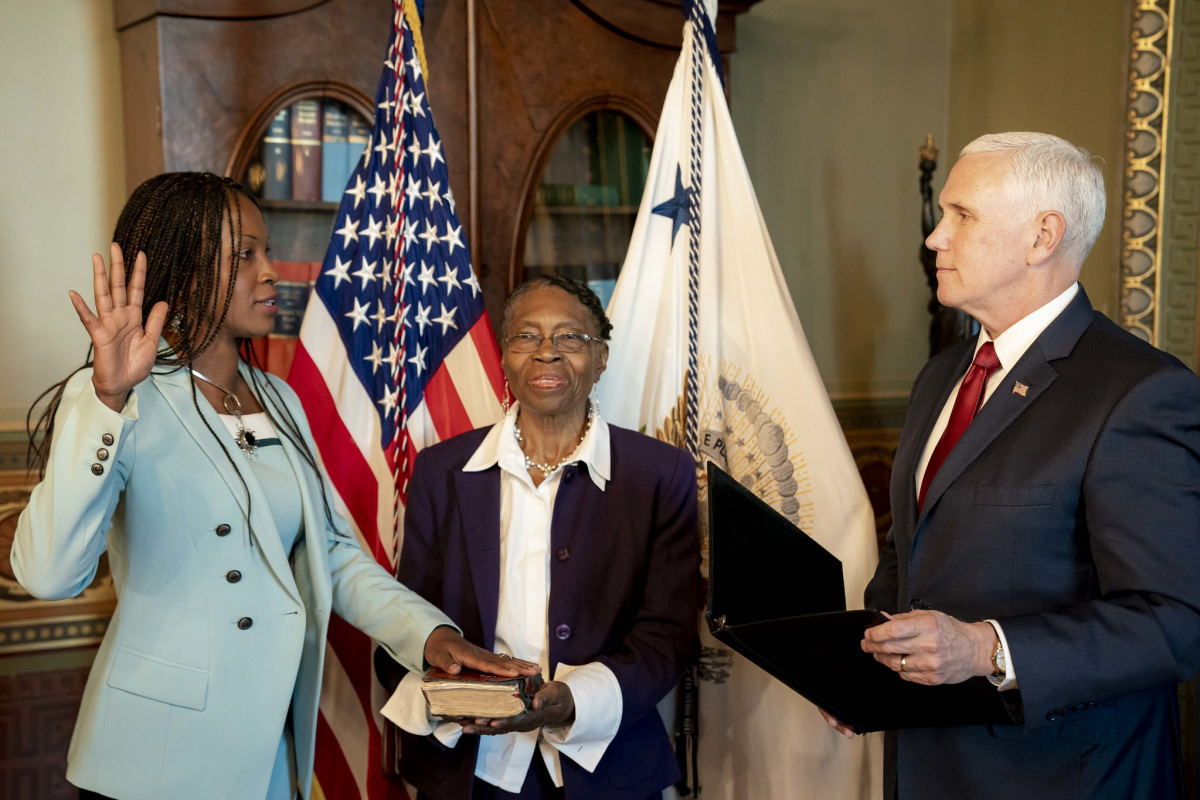 Aurelia Skipwith. an African American woman with long dark hair and wearing a light blue suit, holds one hand in the air and another on a book held by an older African American woman, and takes an oath from Vice President Pence, a white man with gray hair and wearing a dark suit.