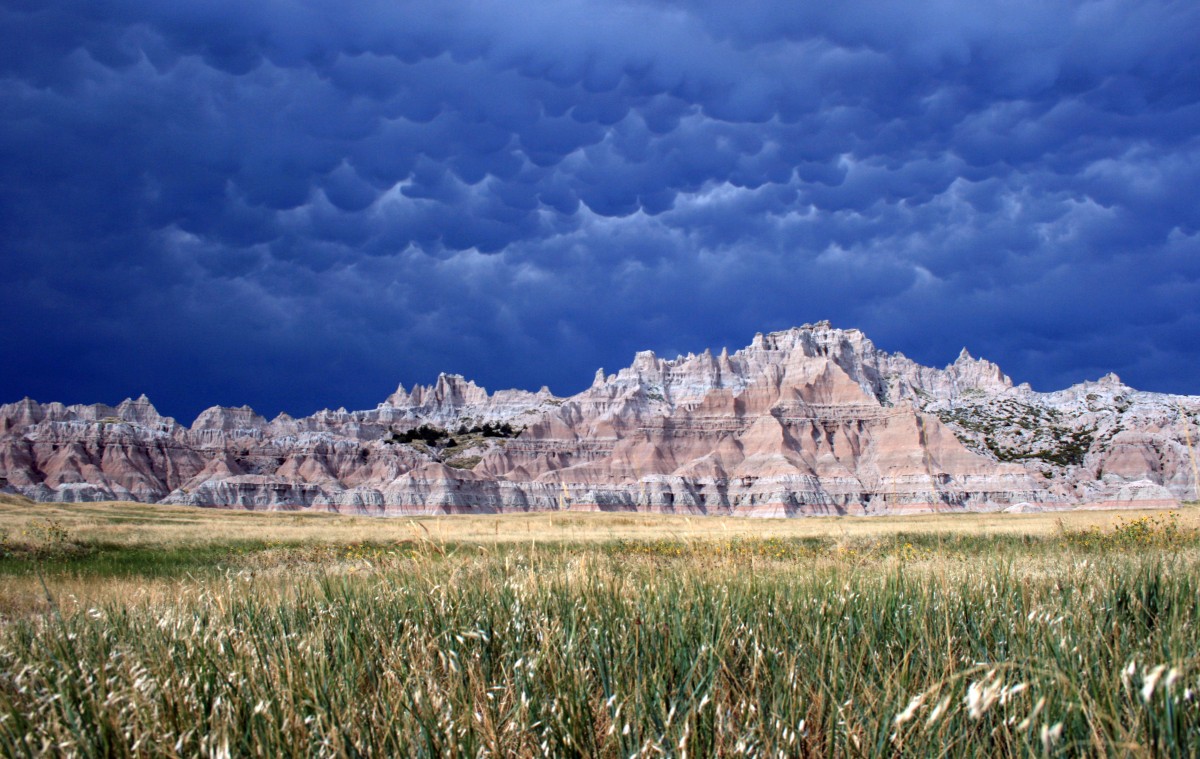 Jagged, striped mountains tower above tall grassy plains beneath dark gray and blue rolling storm clouds.