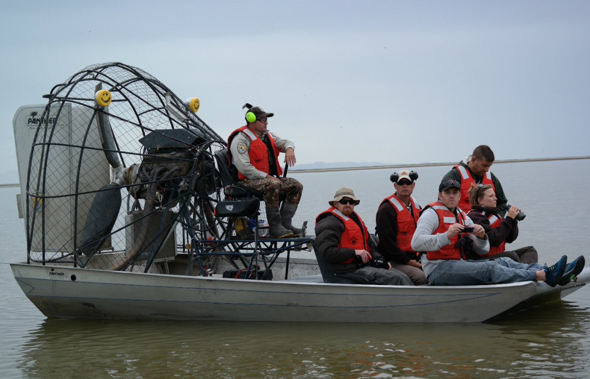 Six people wearing life preservers sit on an airboat floating on calm water.