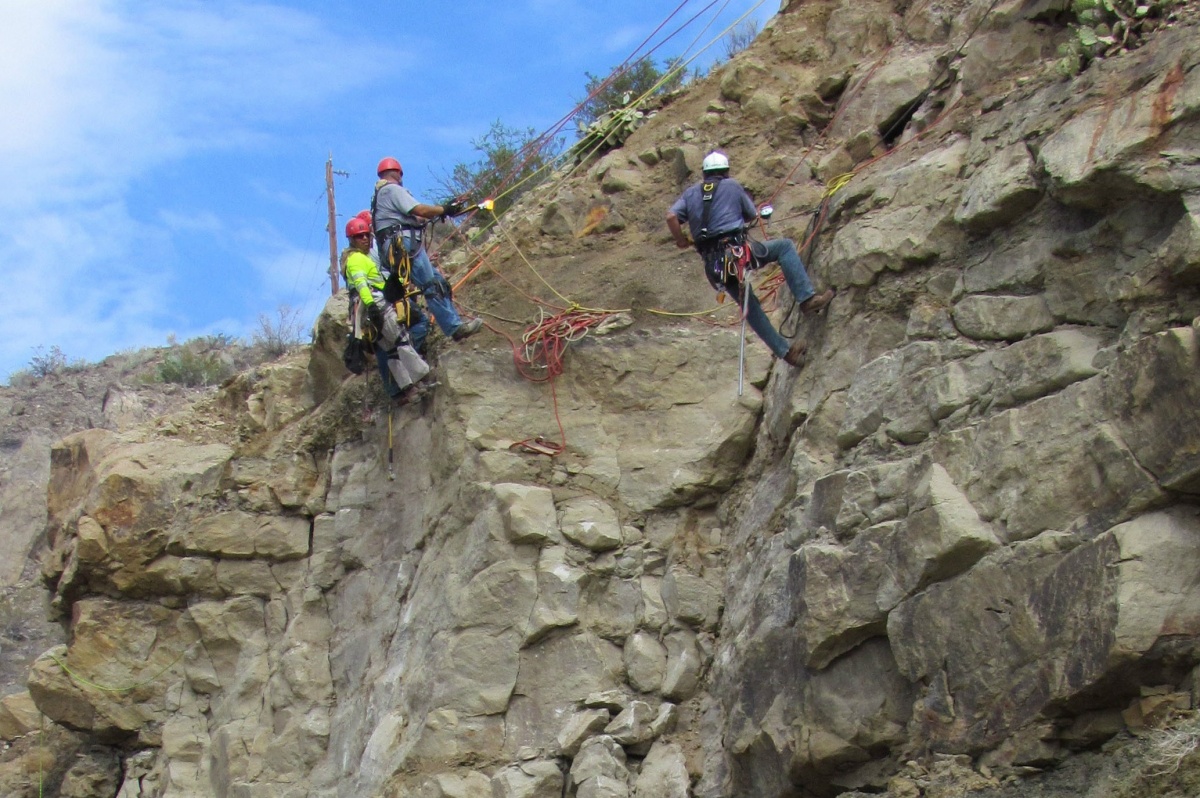 Three men in climbing gear and helmets hangs by ropes on the face of a rocky cliff.