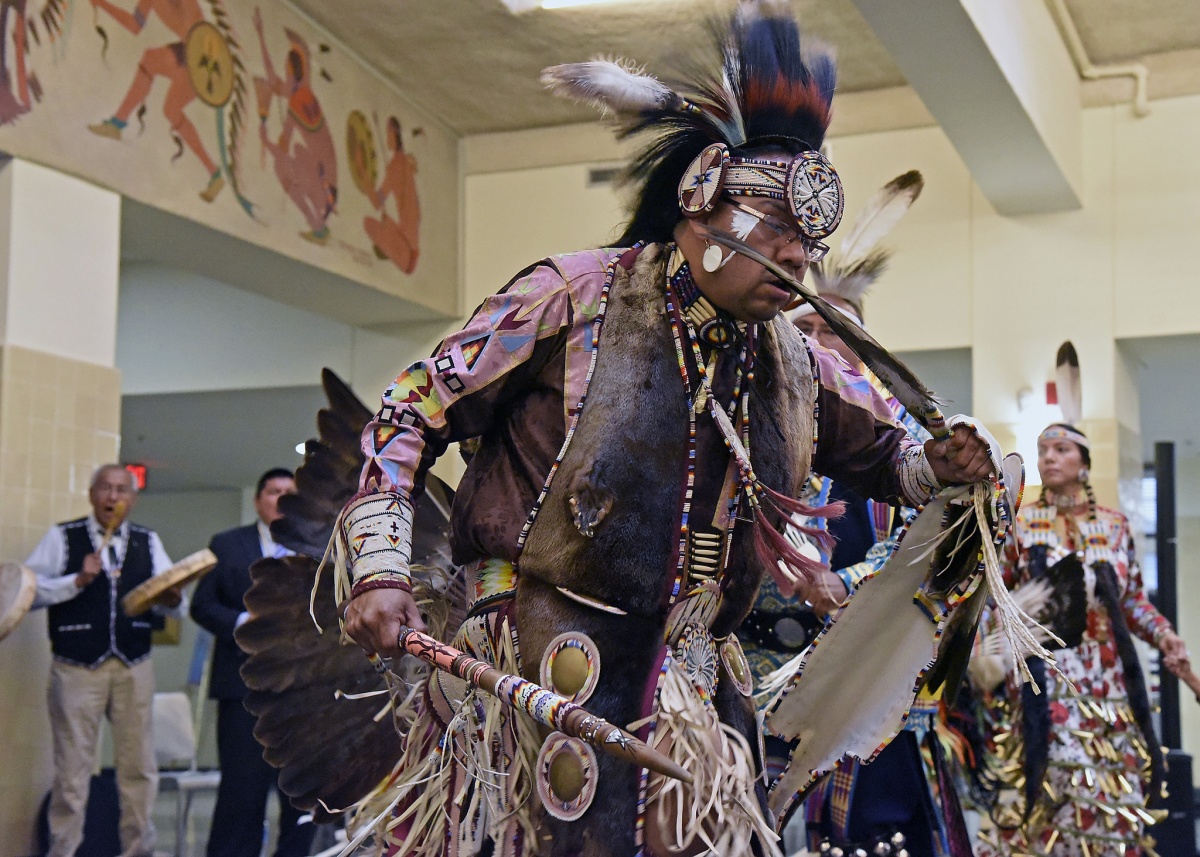 A Native American man wearing traditional clothes and feathers dances inside a large room.
