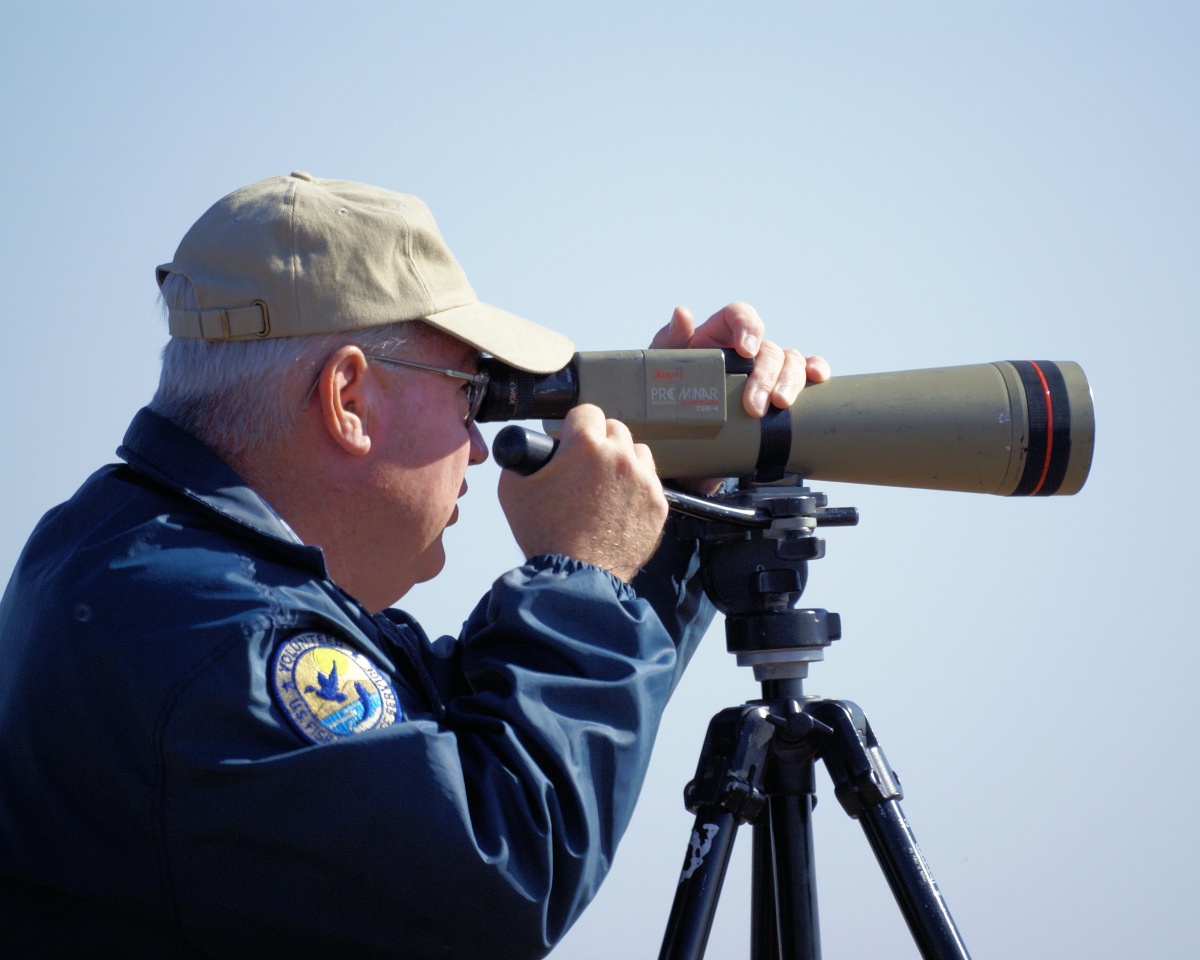 A man in a blue jacket and tan hat looks through a telescope.