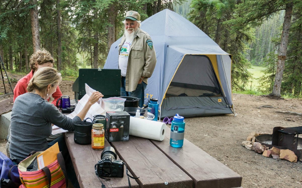 A bearded campground host stands in front of two people sitting on a picnic table looking at a map.