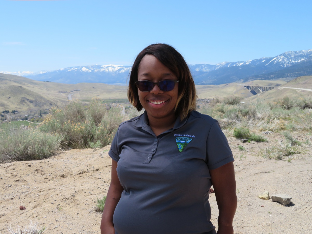 Kemba Anderson, an African American woman wearing sunglasses and a gray BLM shirt, stands on a sandy hilltop with mountains in the distance behind her.