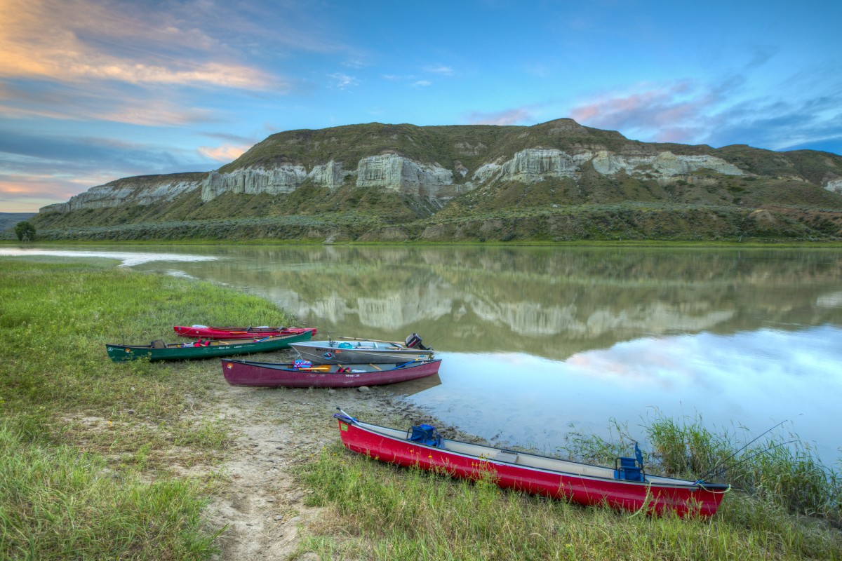 Five colorful canoes are pulled up on the grassy bank of a wide, calm river that has a high rocky cliff rising on the opposite side.