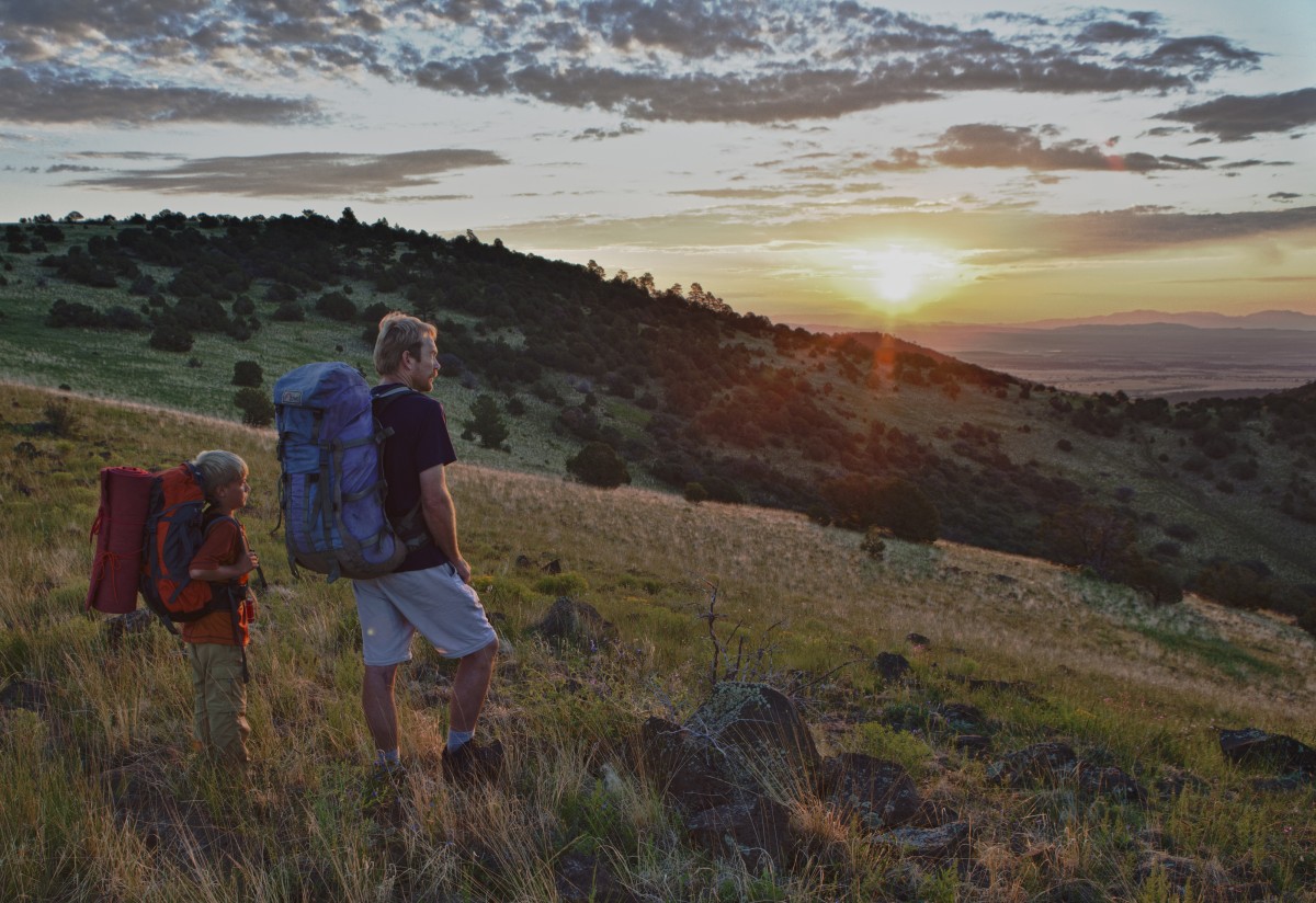 Adult and young child with large backpacks gaze across a green mountain at a distant sunset.