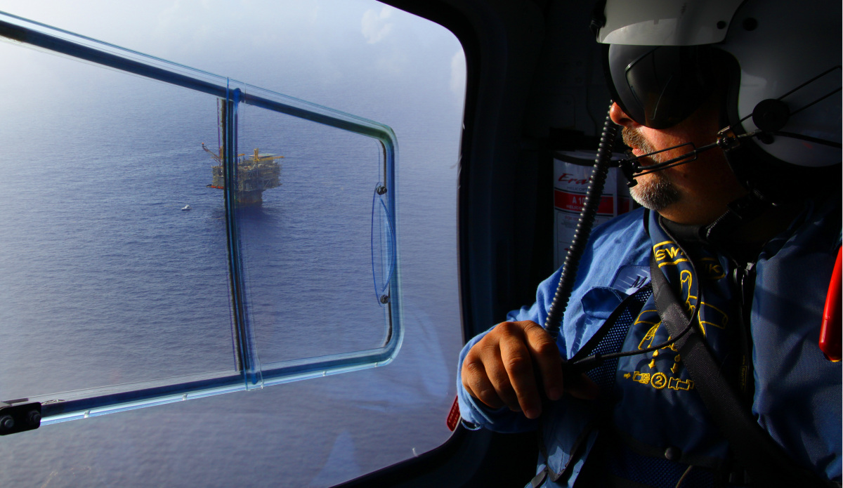 A white man in a flight helmet looks out a helicopter window down at an oil drilling platform on the water below.