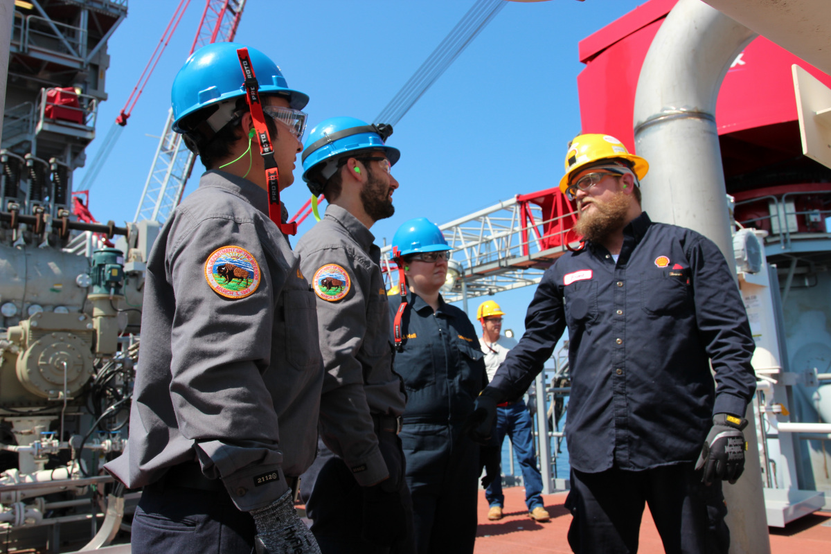 A small group of men and women in work clothes and hard hats talk together while standing on the metal deck of a large drilling platform.