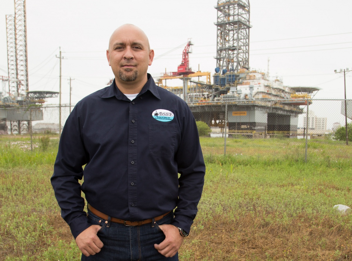 Tommy Treagle, a white man in a navy blue work shirt and jeans, stands in a field with a large metal drilling rig behind him.