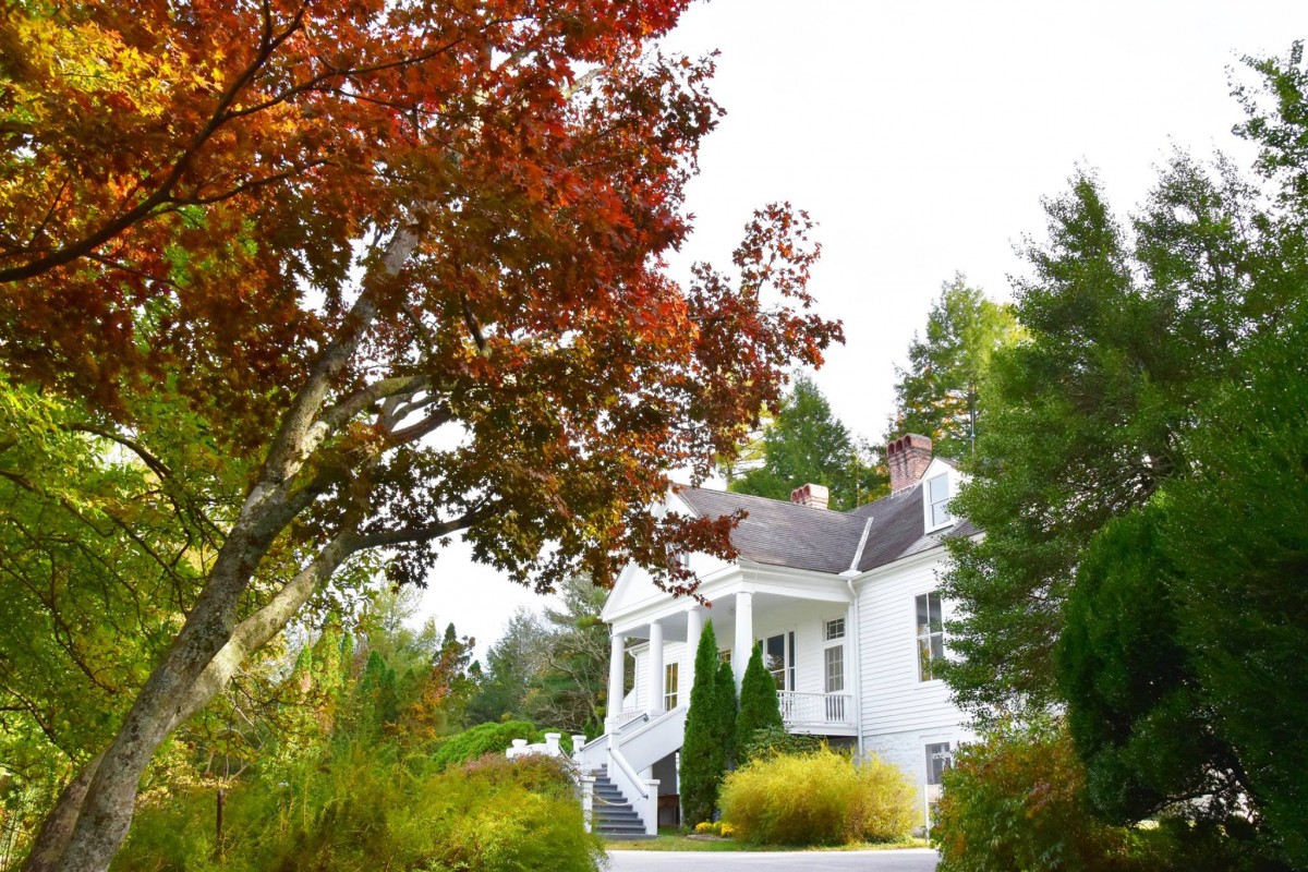 Carl Sandburg's simple white home is visible through autumn leaves and leafy green trees with a pale sky.