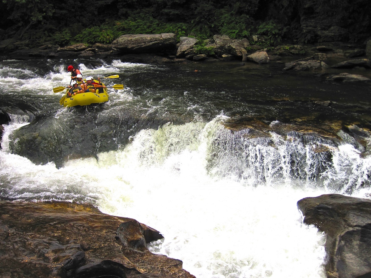 Cinque persone con caschi siedono in un gommone giallo e remano lungo un fiume verso una grande rapida e rocce bianche.