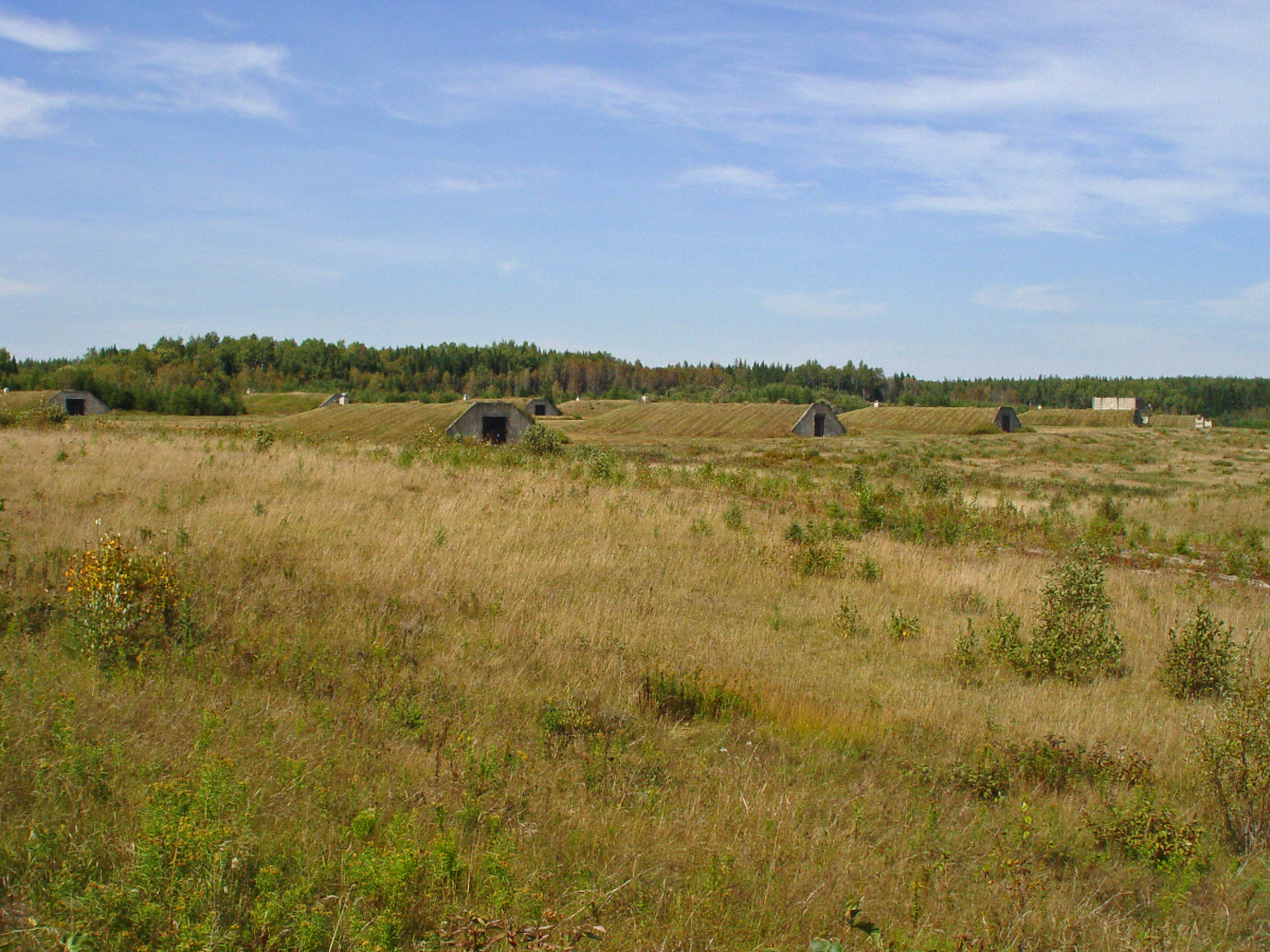 Two lines of low concrete buildings with sod roofs stand in an open field.